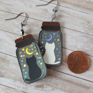 May include: A pair of silver dangle earrings with a black and white cat silhouette inside a glass jar with a crescent moon and stars. The jars are filled with iridescent glitter.