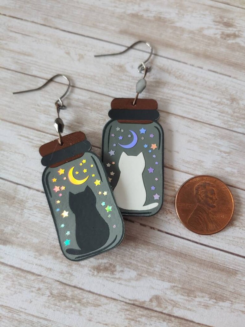 May include: A pair of silver dangle earrings with a black and white cat silhouette inside a glass jar with a crescent moon and stars. The jars are filled with iridescent glitter.