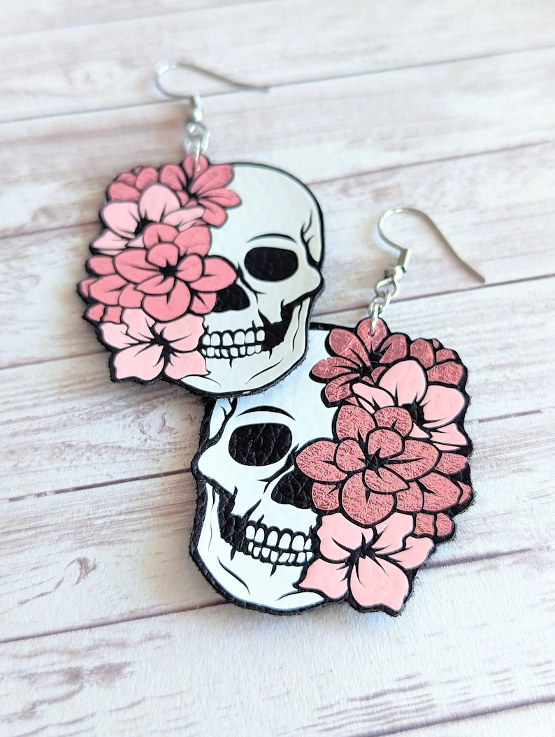 May include: A pair of dangle earrings featuring a white skull with pink flowers covering one eye. The earrings are made of leather and have silver hooks.