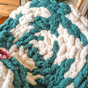 Op de afbeelding: Een handgemaakte, dikke gebreide deken met een spiraalpatroon. De deken heeft afwisselende secties van cr&egrave;me en turquoise garen, wat een aantrekkelijke textuur cre&euml;ert. De deken is gemaakt van dik, zacht garen.