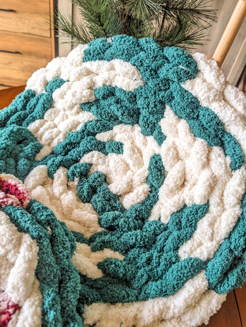 Op de afbeelding: Een handgemaakte, dikke gebreide deken met een spiraalpatroon. De deken heeft afwisselende secties van cr&egrave;me en turquoise garen, wat een aantrekkelijke textuur cre&euml;ert. De deken is gemaakt van dik, zacht garen.