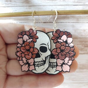 May include: A pair of dangle earrings featuring a white skull with black details and a rose gold floral design. The flowers are pink and rose gold.