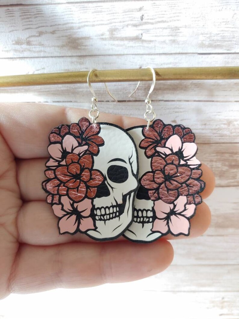 May include: A pair of dangle earrings featuring a white skull with black details and a rose gold floral design. The flowers are pink and rose gold.