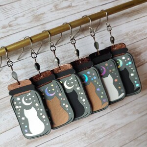 May include: Six pairs of cat-themed earrings, each featuring a cat silhouette inside a glass jar with a crescent moon and stars. The jars are in various colors, including green, brown, and black.