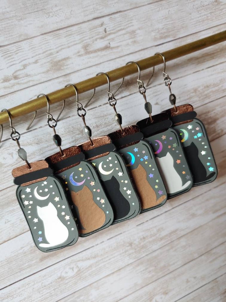 May include: Six pairs of cat-themed earrings, each featuring a cat silhouette inside a glass jar with a crescent moon and stars. The jars are in various colors, including green, brown, and black.