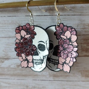 May include: A pair of dangle earrings featuring a white skull with black details and pink glitter flowers. The earrings are made of leather and have silver hooks.