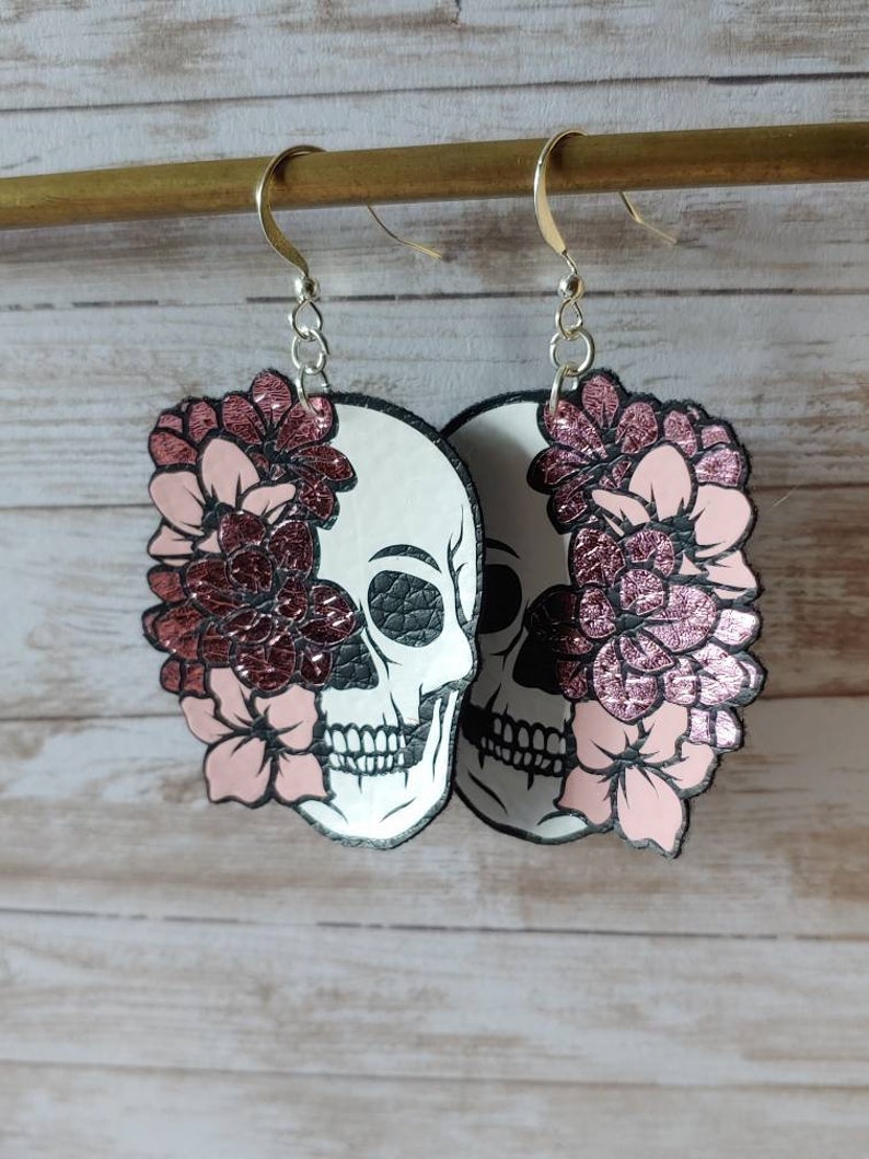 May include: A pair of dangle earrings featuring a white skull with black details and pink glitter flowers. The earrings are made of leather and have silver hooks.