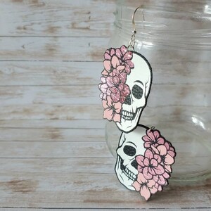 May include: A pair of dangle earrings featuring a white skull with pink flowers on each side. The skull is outlined in black and has a textured surface. The flowers are a light pink color and have a glitter finish.