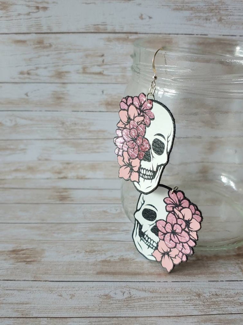 May include: A pair of dangle earrings featuring a white skull with pink flowers on each side. The skull is outlined in black and has a textured surface. The flowers are a light pink color and have a glitter finish.