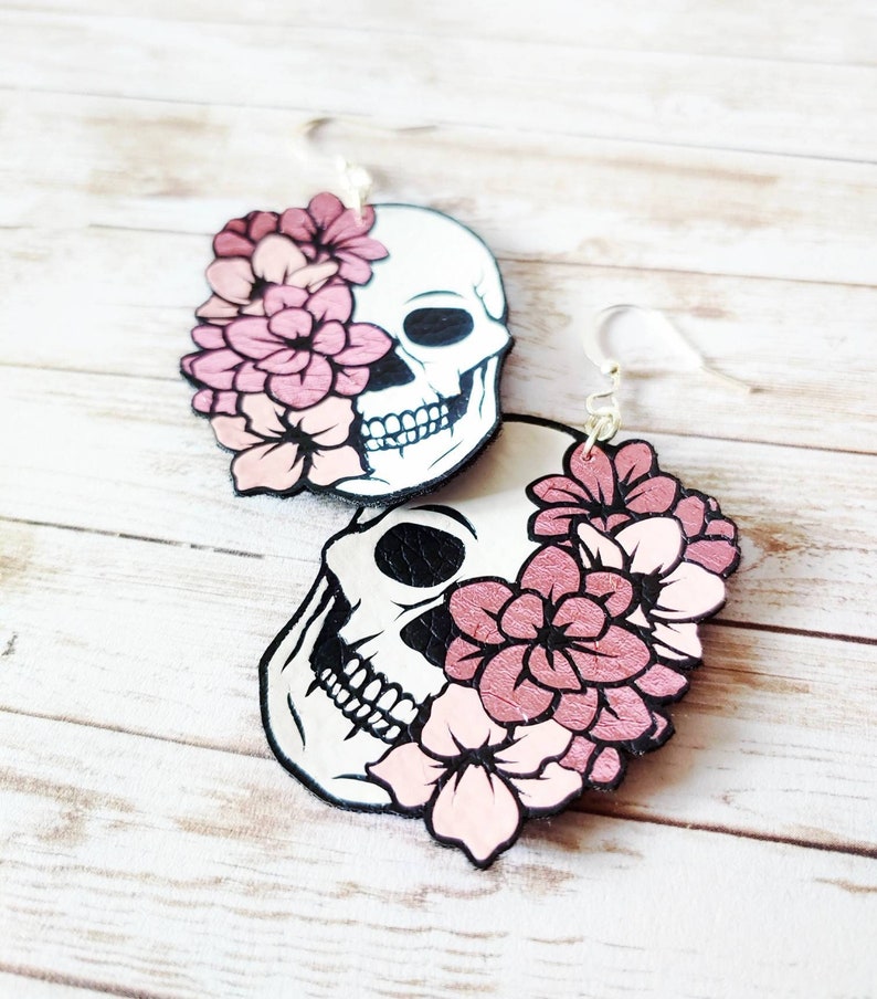May include: A pair of dangle earrings featuring a white skull with black details and pink flowers. The skull is partially covered by the flowers.
