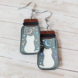 May include: A pair of dangle earrings featuring a white cat silhouette inside a glass jar with a crescent moon and stars. The jar is painted in shades of gray and brown.