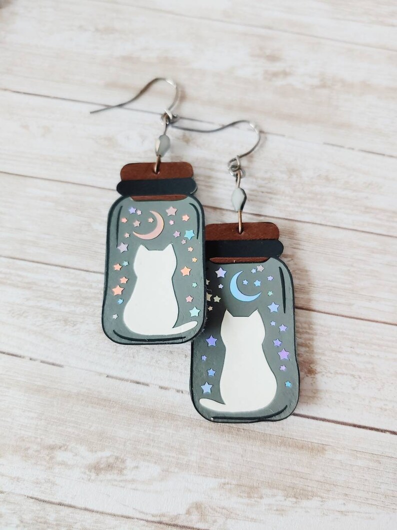 May include: A pair of dangle earrings featuring a white cat silhouette inside a glass jar with a crescent moon and stars. The jar is painted in shades of gray and brown.