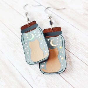 May include: A pair of dangle earrings featuring a cat inside a glass jar with a crescent moon and stars. The jar is painted in a light blue color with a brown lid. The cat is brown and the stars are iridescent.