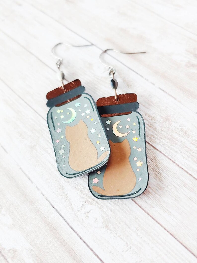 May include: A pair of dangle earrings featuring a cat inside a glass jar with a crescent moon and stars. The jar is painted in a light blue color with a brown lid. The cat is brown and the stars are iridescent.