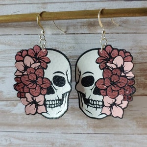 May include: A pair of dangle earrings featuring a white skull with black details and a pink and rose gold floral design.