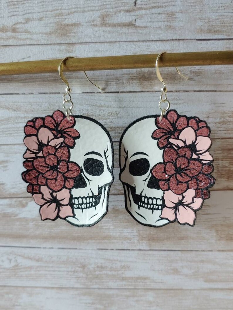 May include: A pair of dangle earrings featuring a white skull with black details and a pink and rose gold floral design.
