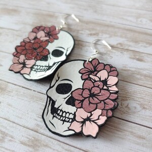 May include: A pair of dangle earrings featuring a white skull with pink and red flowers covering one eye. The earrings have silver hooks.