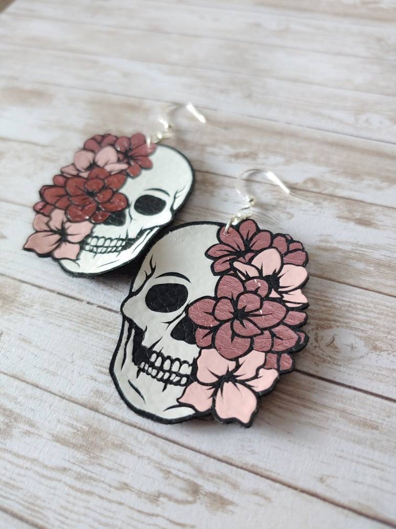 May include: A pair of dangle earrings featuring a white skull with pink and red flowers covering one eye. The earrings have silver hooks.