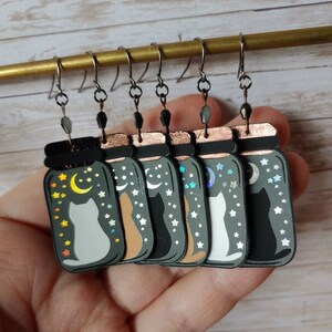 May include: Six pairs of dangle earrings featuring a cat silhouette inside a glass jar with a crescent moon and stars. The jars are in various colors, including black, white, brown, and gray.