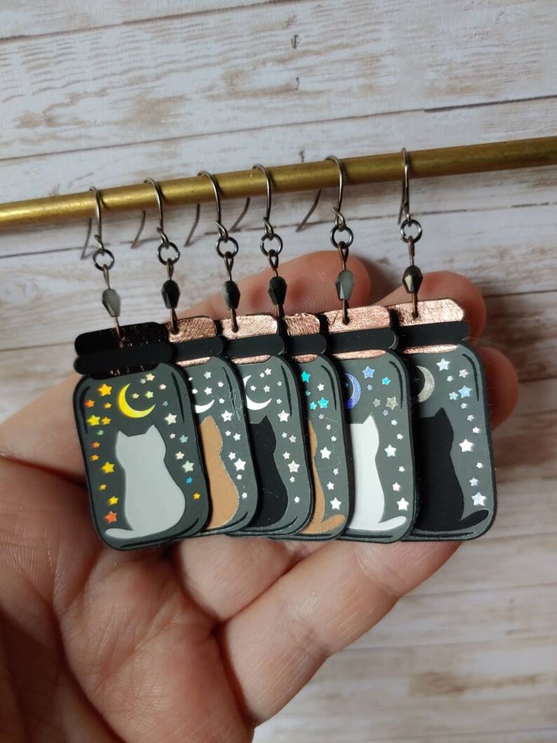 May include: Six pairs of dangle earrings featuring a cat silhouette inside a glass jar with a crescent moon and stars. The jars are in various colors, including black, white, brown, and gray.