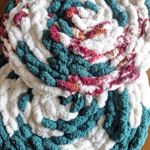 Op de afbeelding: Close-up van twee handgemaakte, cirkelvormige, dik gebreide items. De items hebben een spiraalvormig ontwerp met afwisselende secties van wit, teal en secties met roze en oranje tinten. De textuur is zacht en pluche, wat duidt op een gezellig woondecoratie-item.