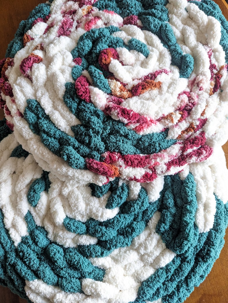 Op de afbeelding: Close-up van twee handgemaakte, cirkelvormige, dik gebreide items. De items hebben een spiraalvormig ontwerp met afwisselende secties van wit, teal en secties met roze en oranje tinten. De textuur is zacht en pluche, wat duidt op een gezellig woondecoratie-item.