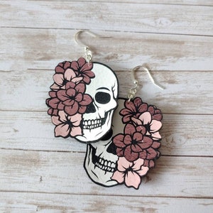 May include: A pair of wooden earrings featuring a skull design with pink flowers. The earrings are a light brown color and have silver hooks.