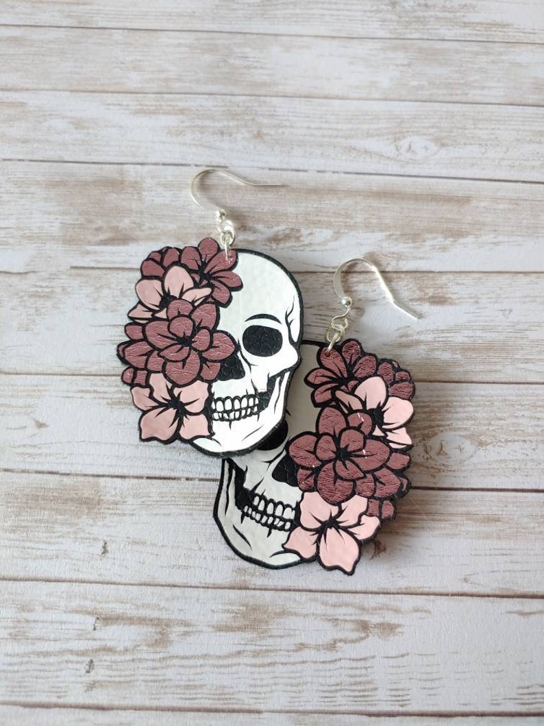May include: A pair of wooden earrings featuring a skull design with pink flowers. The earrings are a light brown color and have silver hooks.
