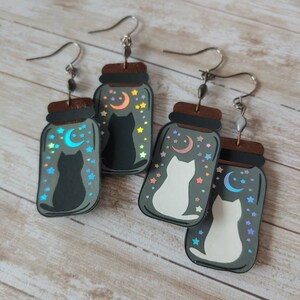 May include: A pair of cat-themed earrings with a black cat and a white cat inside a glass jar with a crescent moon and stars. The jars are made of wood and have a holographic finish.