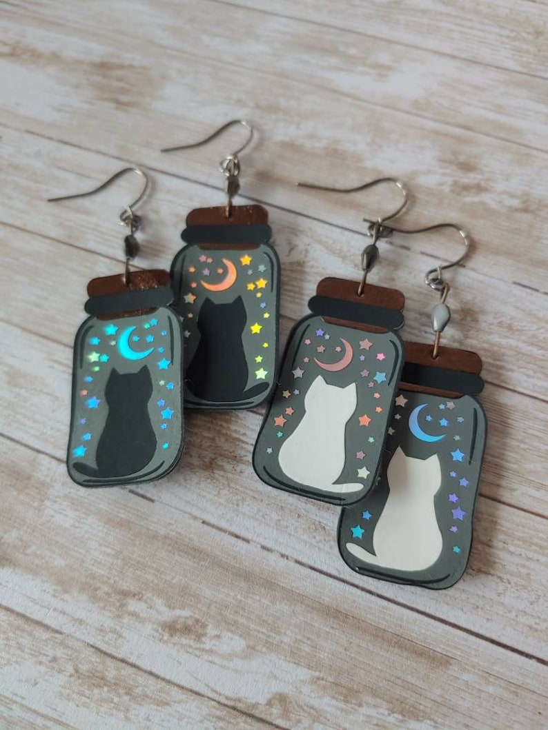 May include: A pair of cat-themed earrings with a black cat and a white cat inside a glass jar with a crescent moon and stars. The jars are made of wood and have a holographic finish.