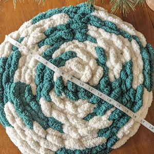 Op de afbeelding: Een ronde, handgebreide vloerkleed met een spiraalpatroon, bestaande uit afwisselende secties van dikke turquoise en witte garen. De vloerkleed heeft een diameter van ongeveer 46 cm. De textuur van het garen is zichtbaar, wat een gezellige uitstraling cre&euml;ert.