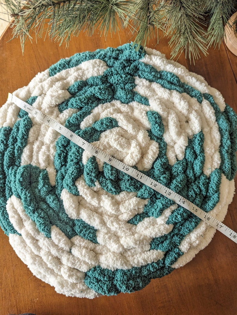 Op de afbeelding: Een ronde, handgebreide vloerkleed met een spiraalpatroon, bestaande uit afwisselende secties van dikke turquoise en witte garen. De vloerkleed heeft een diameter van ongeveer 46 cm. De textuur van het garen is zichtbaar, wat een gezellige uitstraling cre&euml;ert.