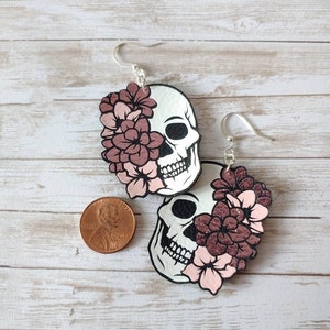 May include: A pair of silver skull earrings with pink flowers. The earrings are about the size of a US penny.