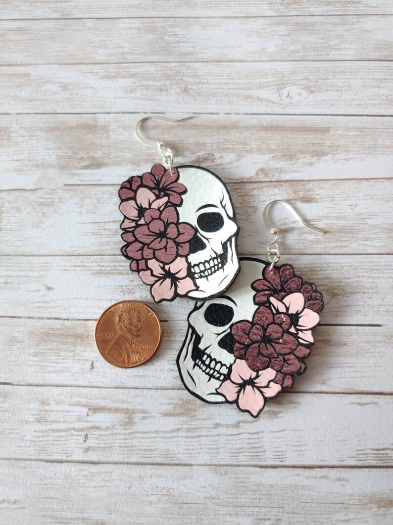 May include: A pair of silver skull earrings with pink flowers. The earrings are about the size of a US penny.