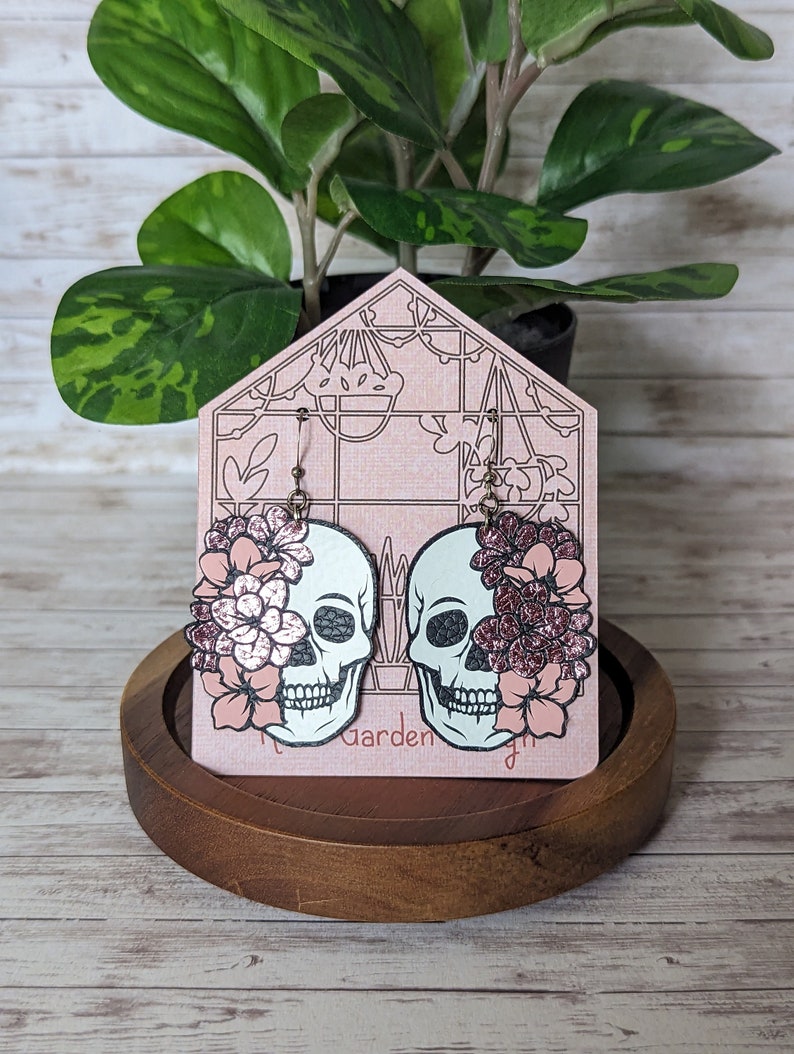 May include: A pair of dangle earrings featuring a black and white skull design with pink glitter flowers on each side. The earrings are displayed on a pink and white card with the text "The Garden".
