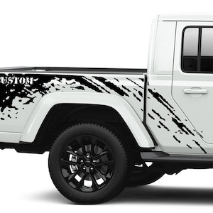 May include: A white Jeep Gladiator pickup truck with black mud-splatter graphics on the side of the truck bed. The graphics include the word "CUSTOM" in black lettering.