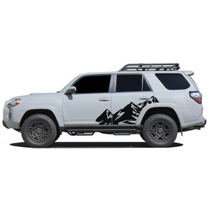 May include: A white SUV with a black mountain decal on the side. The vehicle has a roof rack and black wheels.
