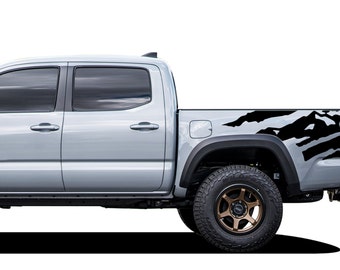Puede incluir: Una camioneta pick-up plateada con gráficos laterales negros. La camioneta tiene una caja de carga negra y un parachoques trasero negro. La camioneta está sobre un fondo blanco.