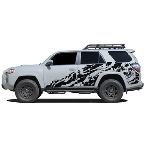 May include: A white SUV with black skull and tyre tread graphics on the side. The vehicle has a roof rack and black wheels.