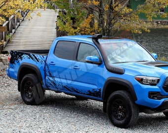Puede incluir: Una camioneta pickup azul brillante con detalles en negro y un snorkel. La camioneta tiene ruedas y neumáticos negros, y un diseño gráfico negro en el lateral. Está estacionada sobre una superficie de grava cerca de un puente de madera.