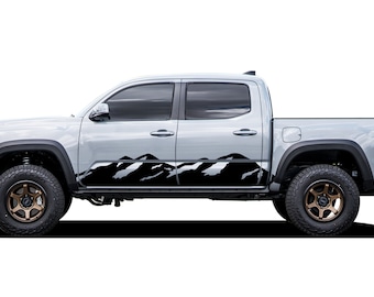 Puede incluir: Una camioneta plateada con calcomanías de montañas negras en el lateral. La camioneta tiene ruedas negras con llantas de bronce.