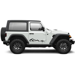 Op de afbeelding: Een witte Jeep Wrangler met zwarte bergdecals aan de zijkant. De Jeep heeft zwarte wielen en een zwart hardtop.
