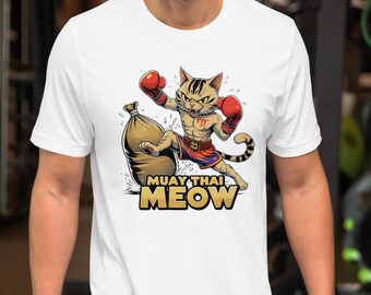 Puede incluir: Camiseta blanca con un gato de dibujos animados con equipo de boxeo Muay Thai, incluyendo guantes y pantalones cortos rojos, pateando un saco de arena. El texto "MUAY THAI MEOW" está impreso debajo en amarillo y negro.
