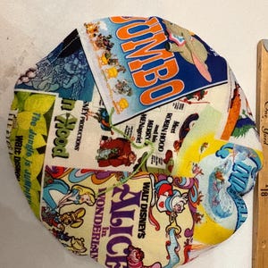 Op de afbeelding: Een rond, veelkleurig stoffen item met verschillende Disney-filmposters, waaronder "Dumbo", "Alice in Wonderland" en "Robin Hood". Ernaast ligt een houten liniaal met inch-maten.
