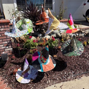 May include: A collection of six fabric witch hats displayed on metal stakes. Each hat features a unique patchwork design with various colorful patterns, including polka dots, floral prints, and geometric shapes in shades of orange, green, pink, and black.