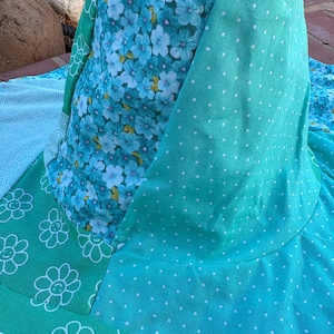 May include: A patchwork textile featuring various floral and polka dot patterns in shades of blue and green. The fabric includes a floral print with white flowers, a teal fabric with white polka dots, and a green fabric with white smiling flower outlines.