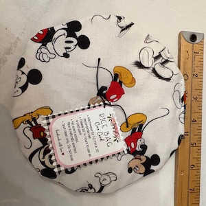 May include: A white rice bag with a Mickey Mouse pattern, featuring black, red, and yellow details. A wooden ruler is next to the bag. The bag has a tag with care instructions.