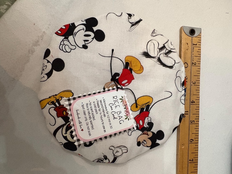 Op de afbeelding: Een witte rijstzak met een Mickey Mouse-patroon, met zwarte, rode en gele details. Een houten liniaal staat naast de zak. De zak heeft een label met onderhoudsinstructies.