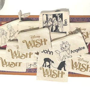 May include: A collection of off-white canvas zip pouches featuring various Disney-themed graphics and text. Designs include the Disney Wish logo in gold glitter, character illustrations like Marie from The Aristocats, and personalised names.
