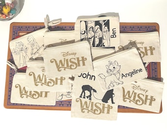 May include: A collection of off-white canvas zip pouches featuring various Disney-themed graphics and text. Designs include the Disney Wish logo in gold glitter, character illustrations like Marie from The Aristocats, and personalised names.